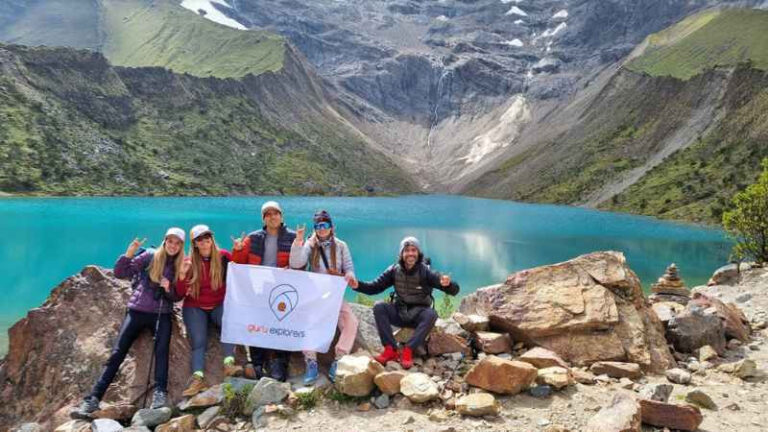 Grupo de viajeros con bandera de Guru Explorers en la Laguna Humantay con aguas turquesas y el nevado Salkantay al fondo