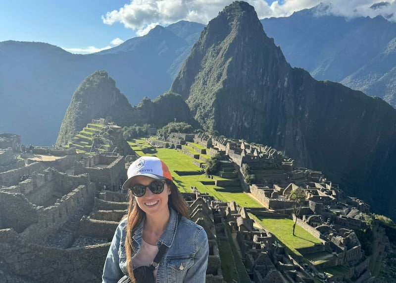Viajante com boné da Guru Explorers sorrindo em Machu Picchu, com a cidadela inca e a montanha ao fundo